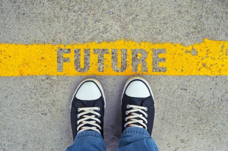 Looking down at a pair of trainers standing on a road, straddling a painted yellow line stencilled with the word FUTURE.