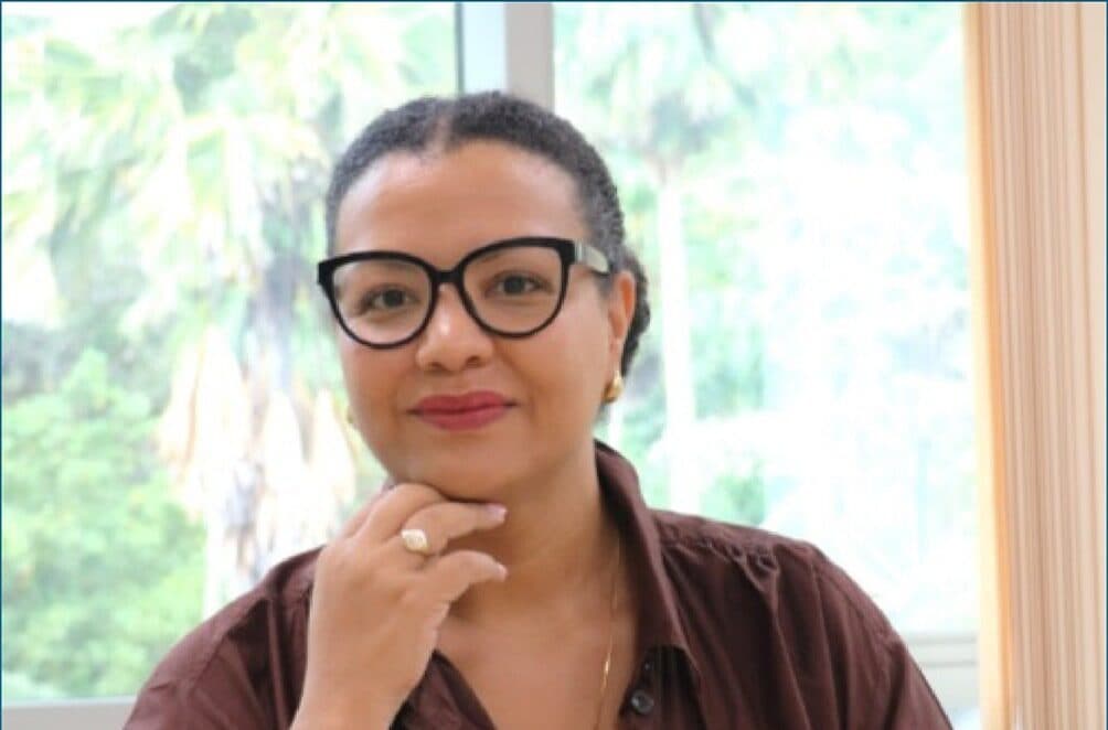 Minister Amanda Bernstein seated, resting her chin on one hand, photographed against a bright garden window.