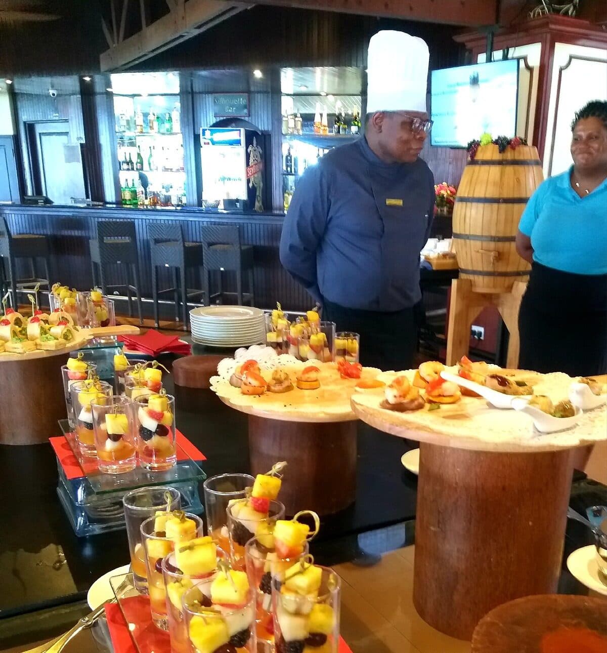 A chef in whites beside a buffet display of canapés and desserts in wooden serving vessels.