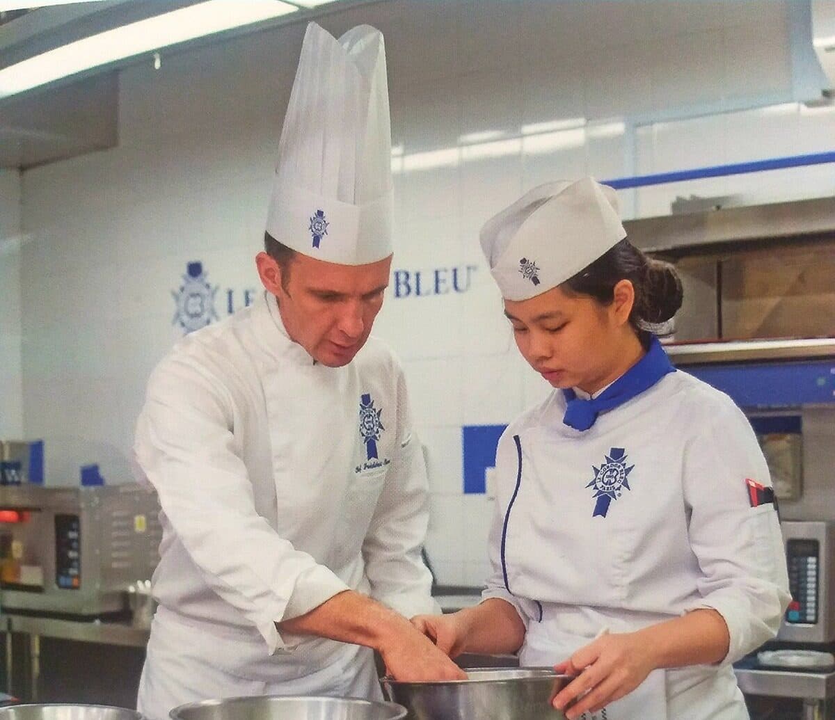 A chef instructor in whites guiding a student's hands over a mixing bowl in a professional teaching kitchen.