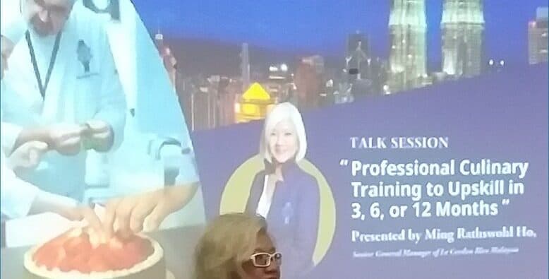 A presentation slide advertising a talk session on professional culinary training, shown on screen above a demonstration counter.