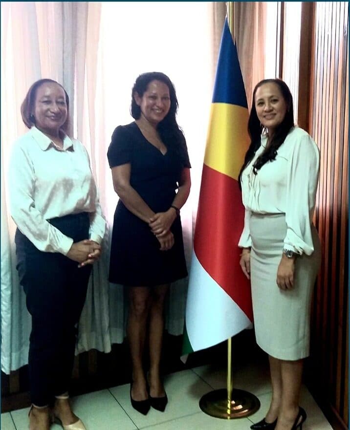 Three women standing beside the Seychelles national flag in a ministerial office.