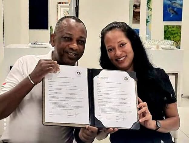 Peter Sinon and Shamita Palit standing side by side holding open the signed memorandum of understanding towards the camera.