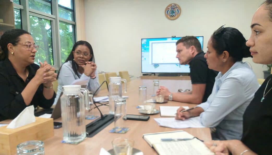 Six people seated around a boardroom table with water glasses and papers, a wall-mounted screen behind them.