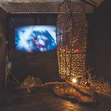A darkened gallery space with a suspended woven fish trap lit from within, beside a projected video work.