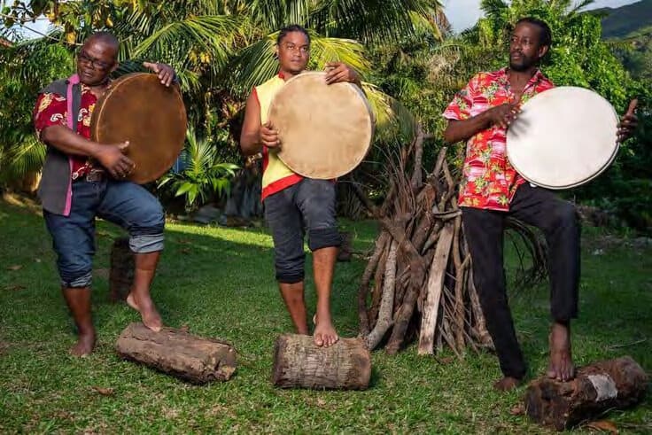 Three Seychellois musicians playing large hand drums outdoors, seated on logs in front of palms and a driftwood structure.